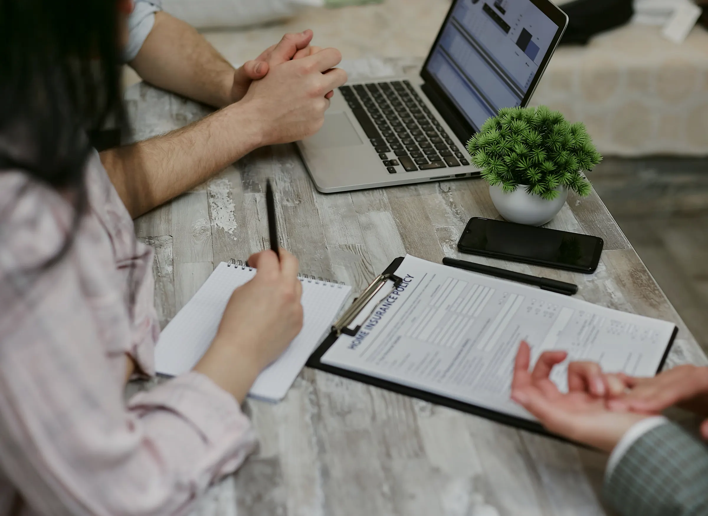 Two people at a desk going over an insurance form on a clipboard, a laptop open beside them
